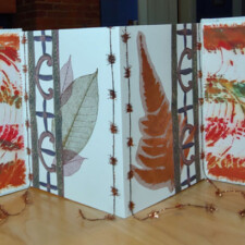 DEBBIE SHANNON
The art seed items I received from Susan Grant were all very powerful visually. I felt that if I put all of them into the same plane it would have been overwhelming. Of course the obvious format for me was a book since that’s a form that I’ve been working in for a long time. An accordion book can be displayed open providing for multiple planes that could highlight some of the work while having an overall design. I made use of everything she gave me without really altering the materials. I added cardboard, white cover paper, inside accordion pages and two small buttons on the front cover. There are actual pieces of trim on the front and back covers. For the inside pages, I used copies of the trim.