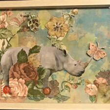 SUE SOBEL “Ambiguous Loss”
A rhinoceros on wheels in an English garden. Ambiguous, puzzling?
This shadow box was influenced by the psychological term ambiguous loss, coined by Dr. Pauline Boss in the 1970’s. It is a loss without closure or a clear understanding which complicates the grieving process. The Pandemic has left many people grieving for the loss of loved ones as we are denied the rituals of visitation and memorials. Covid 19 has caused a personal loss for my family as my husband’s 95 year old mother is isolated and depressed in her senior living facility due to CDC regulations and we are unable to be with her physically and support her emotionally.
Using the Art Seeds sent to me by my partner, Portia Brown, I cut out the rhinoceros from the 2012 WWF calendar and mounted it on Bristol board. The background was gelli printed using some of the floral fabric. Buttons were attached to the feet of the preposterous rhino as wheels. Where the fabric pieces were cut up and expanded, the gaps were filled in with colored pencils. Two kinds of seed beads added visual interest to the flower. A gold translucent gauze fabric, which I had, was layered to give dimension and an other world feeling. Needing more texture in the garden, fabric scraps from the gelli printing were added and enhanced with gelly roll pens. Foam adhesive circles glued on to the backs of the paper and fabric elements supported the different levels of depth in the shadow box.
The lion lurking behind the foliage in the bottom right hand corner, observes, helpless to do anything for the rhinoceros. Coincidentally, the lion pin represents my husband, a Leo. As a way of dealing with the feelings of ambiguous loss, Dr. Boss suggests having a strong religious and spiritual life to cope with the unknown. The jeweled brooch that is suspended above the garden represents my mother in law’s spirituality and our family’s faith.